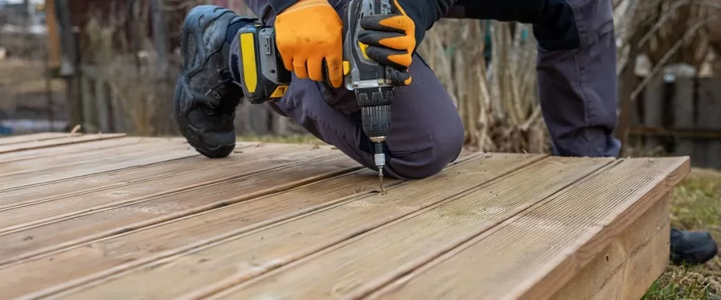 Detailed view of a worker in safety gloves using a cordless drill to secure new wooden boards during a deck replacement project.