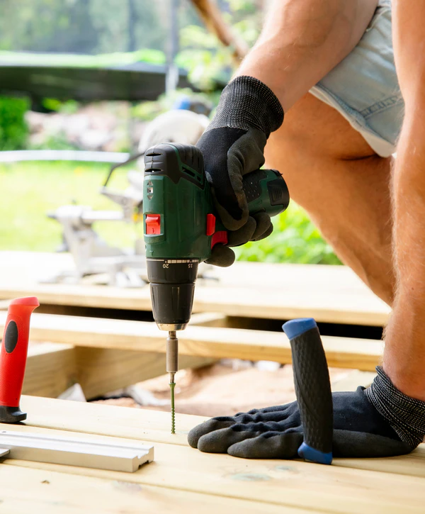 Close-up of a worker representing deck repair companies in Durham, NC, wearing safety gloves and using a cordless drill on a wooden deck subframe.