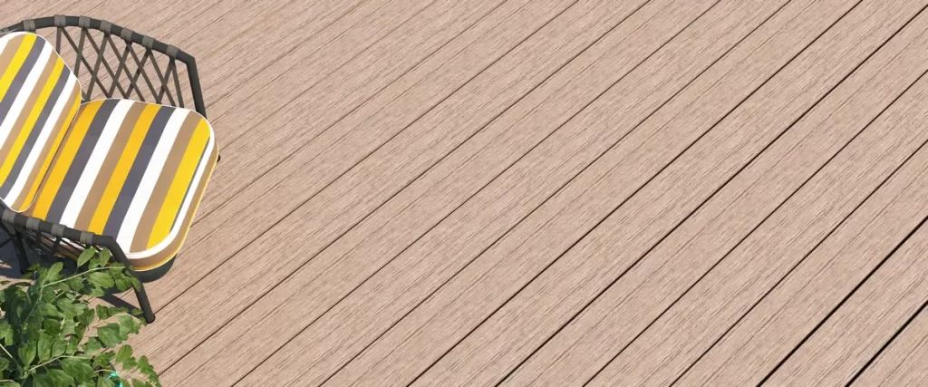 A high-angle close-up shot of light oak-colored composite decking boards featuring a realistic wood grain texture, situated next to a modern striped patio chair and green garden foliage.