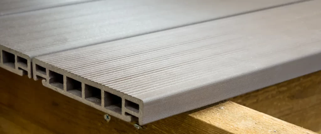An extreme close-up of the cross-section of a grey composite decking board, highlighting the hollow core structure and grooved surface design resting on a wooden joist.