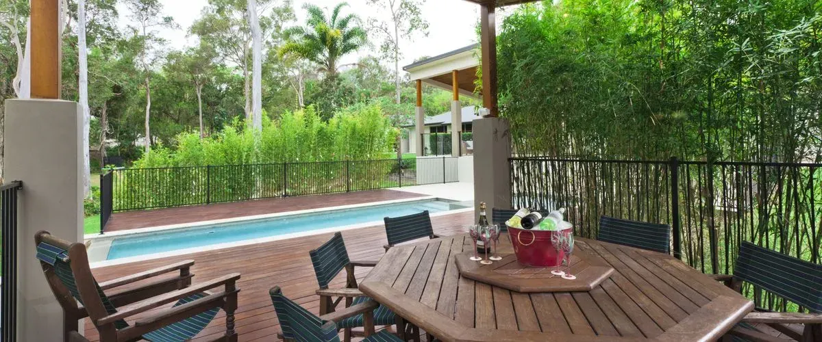 A luxury outdoor living area featuring a large wooden deck surrounding a lap pool with an octagonal wooden dining table and chairs shaded by a lush green bamboo privacy screen.
