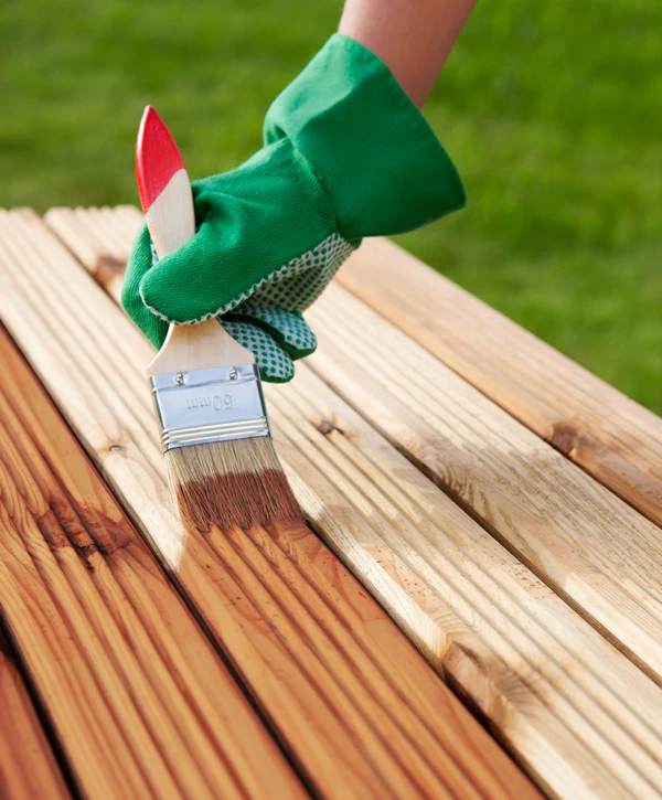 Close-up of a hand in a green safety glove using a 50mm brush to apply protective wood stain to individual planks of a cedar deck.