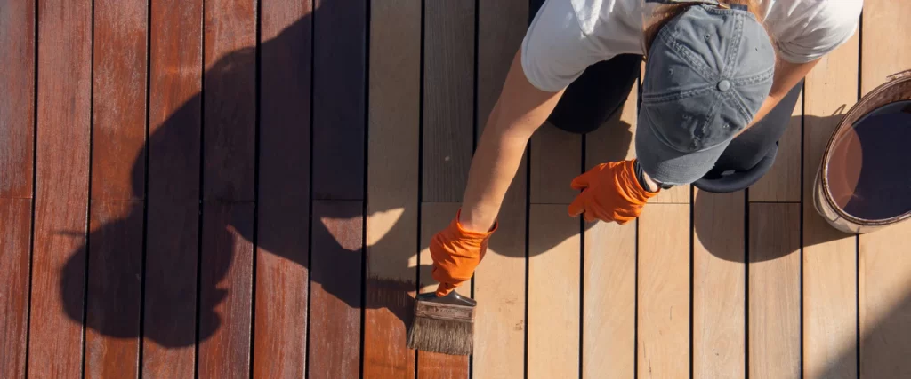 An overhead perspective of a worker in orange gloves applying a dark reddish-brown stain and sealing coat to wooden patio planks with a wide brush.
