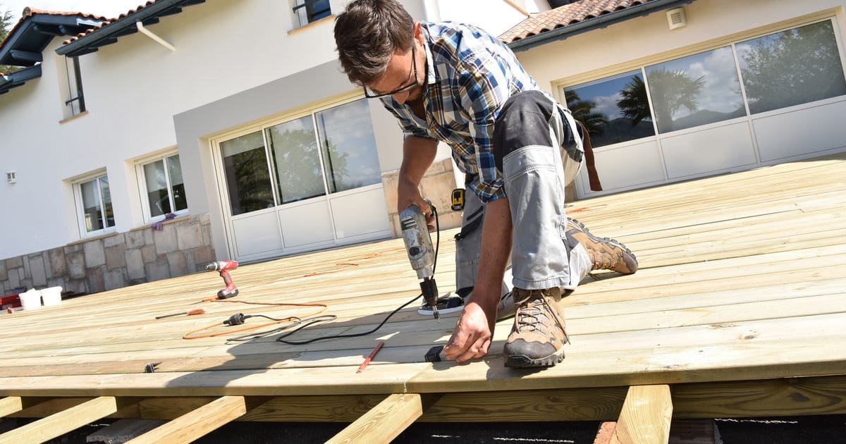 A professional contractor in work gear using a power drill to secure wooden deck boards on a residential property, ensuring the construction meets the latest North Carolina deck building code requirements for safety and stability.