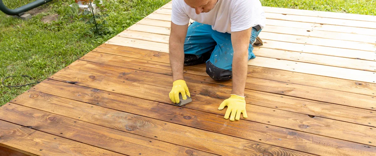 A man wearing yellow gloves kneeling on a wooden deck while applying a rich brown protective wood stain to the individual timber planks using a sanding sponge.
