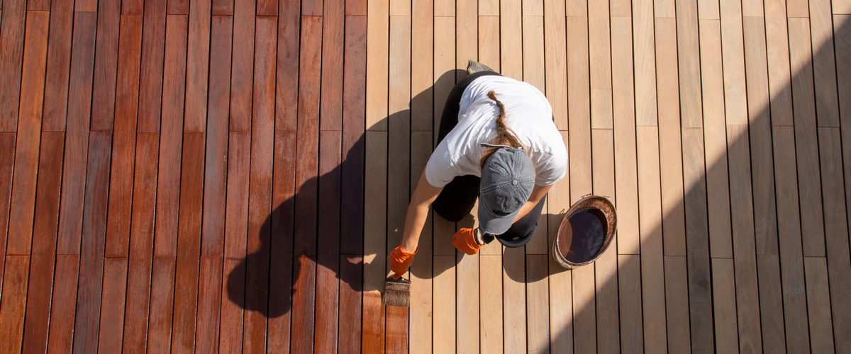 A person wearing a hat and orange gloves kneeling on a wooden deck while applying a rich dark brown stain to the timber planks with a wide brush.