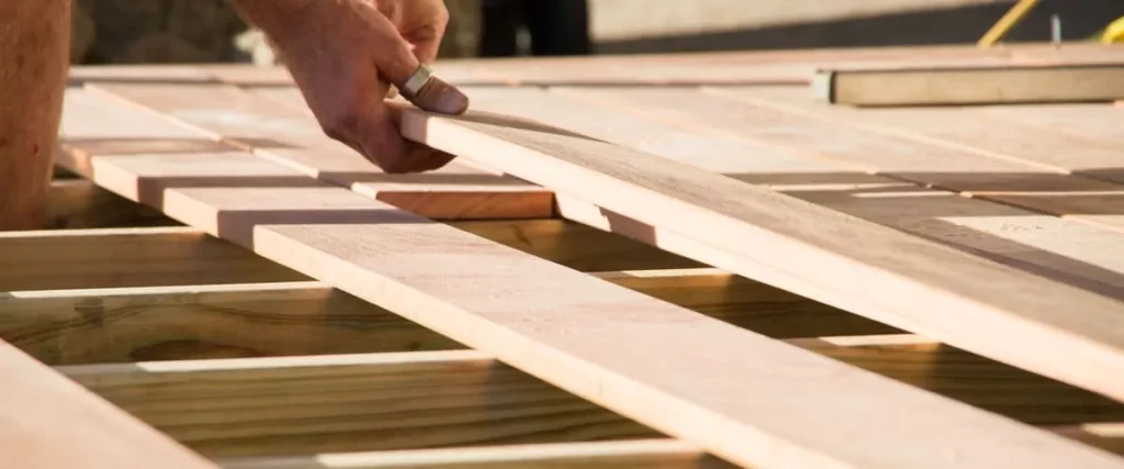 A close-up view of a carpenter's hands carefully aligning a natural timber board onto a wooden deck joist during the construction process.