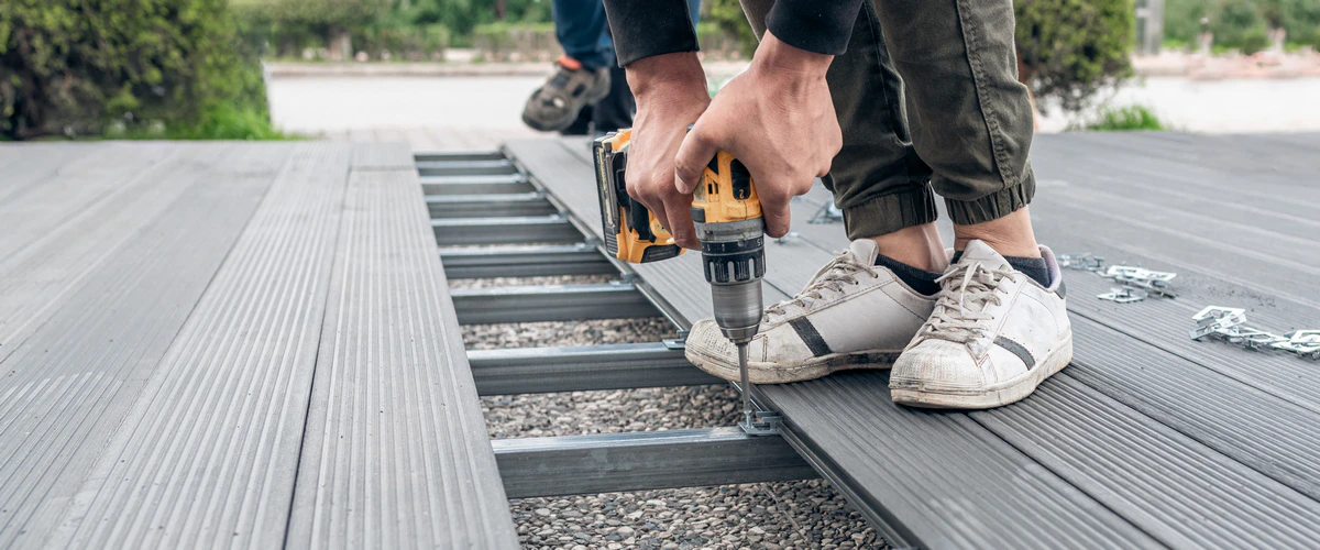 A close-up of a contractor using a yellow power drill to secure grey composite decking boards during an outdoor patio repair project.
