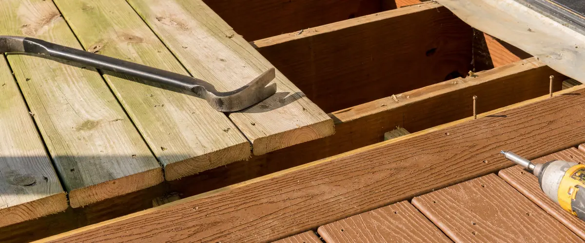 A metal crowbar resting on weathered wooden planks during a home deck repair and demolition project showing the exposed joists.