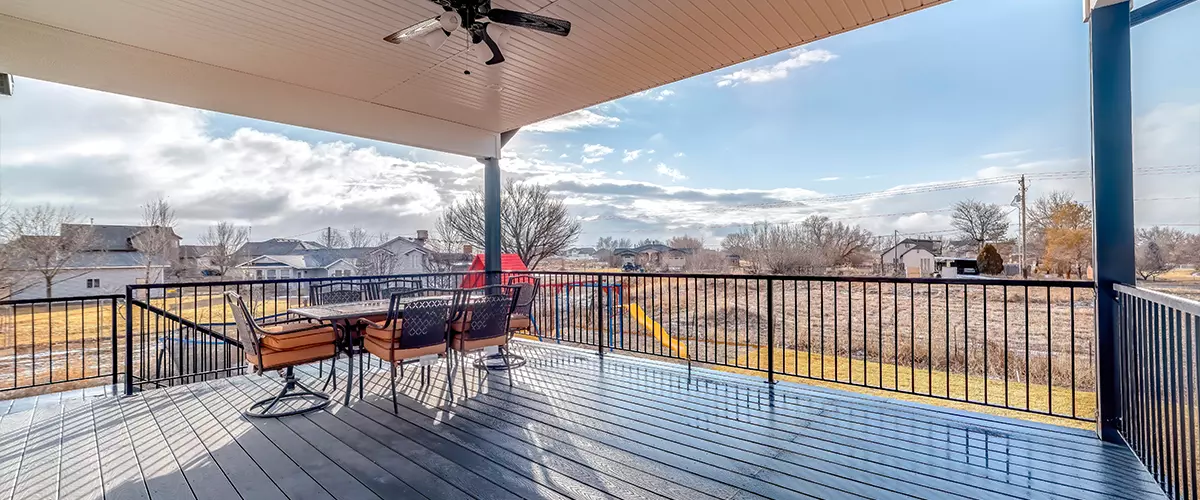 A spacious elevated outdoor deck featuring a dark grey wood floor, a dining table with orange cushioned chairs, and a black metal safety railing overlooking a neighborhood.