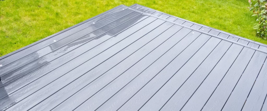 A wide shot of a newly installed grey composite deck with a wet surface showing water resistance, surrounded by a lush green lawn and garden shrubs.
