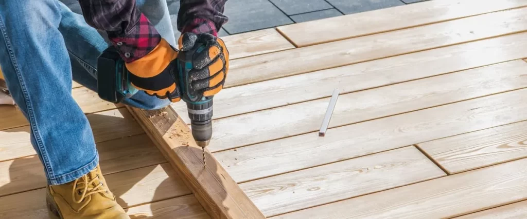 A construction worker wearing safety gloves and boots uses a cordless power drill to bore holes into light-colored wood planks for a backyard deck project.