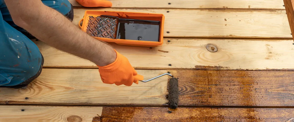 A technician using a paint roller and a tray of dark stain to refinish a wooden deck surface during a deck staining project.