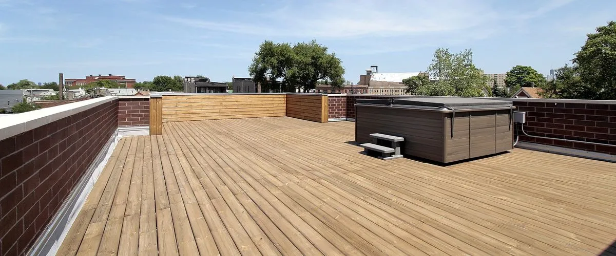 A spacious rooftop wooden deck featuring a built-in hot tub, brick perimeter walls, and a clear view of the city skyline.