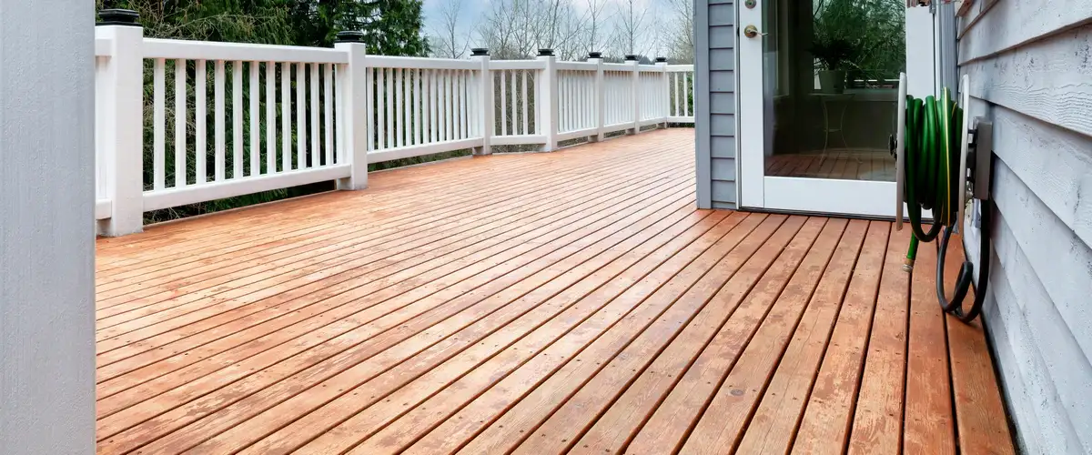 A beautifully finished deck staining project featuring a clean, medium-toned wooden deck next to white safety railings and a coiled green garden hose attached to the house siding.