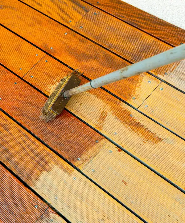 Close up view of a professional from one of the top deck staining companies in Pittsboro, NC, using a stiff brush to apply a rich amber stain to weathered wooden deck boards.